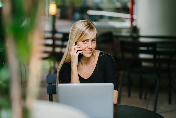 Portrait of a young, attractive and photogenic blond woman talking on her phone with her laptop computer in front of her. She is seated in a cafe during the day as she takes her call. - Foto, immagini