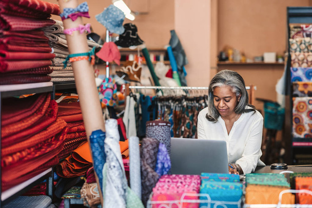 Smiling mature fabric shop owner working on a laptop while standing at a counter surrounded by colorful cloths and textiles  - 写真・画像