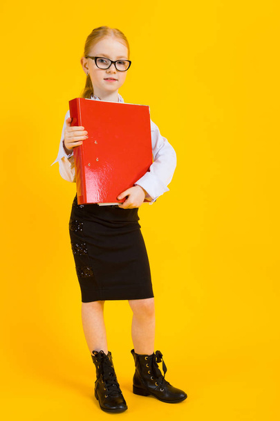 Girl with red hair on a yellow background. A charming girl in transparent glasses is holding a large red folder in her hands. - Photo, image