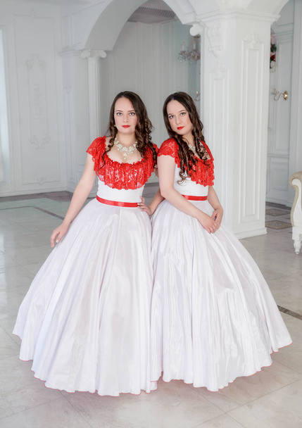 Dos mujeres hermosas en vestidos medievales blancos y rojos con crinolina en la sala
 - Foto, imagen