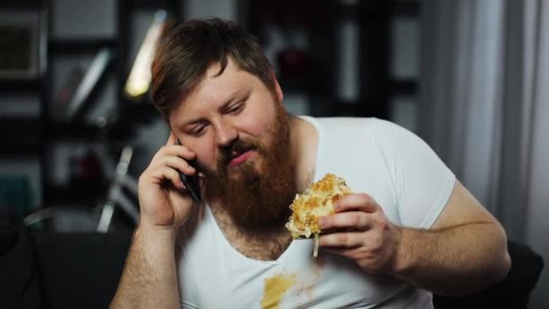 Dirty fat bearded man talks on the smartphone, eating a burger and drinking beer sitting on the couch. Concept of malnutrition, food delivery, communication and obesity - 映像、動画
