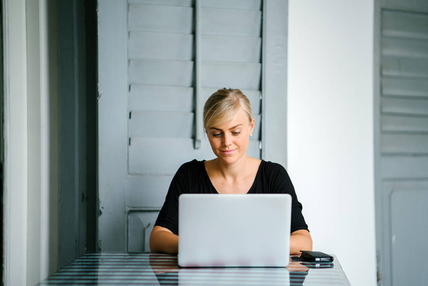 Retrato de una joven y atractiva rubia de startup trabajando en un escritorio durante el día en su portátil. Ella se centra en su trabajo y está escribiendo en su cuaderno y tiene el pelo atado
. - Foto, Imagen
