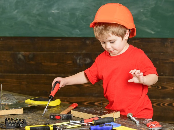 Kid boy holds screwdriver tool. Handcrafting and workshop concept. Child in helmet cute playing as builder or repairer, or handcrafting. Toddler on busy face plays with screwdriver at workshop - Foto, Imagen