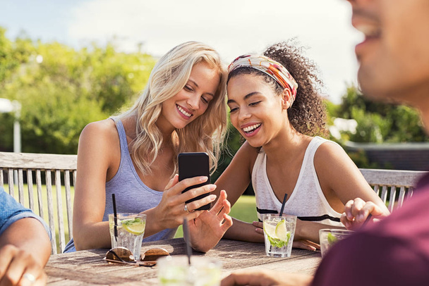 Two young women looking at smart phone and smiling sitting at table outdoor. Happy multiethnic girls taking selfie at outdoor cafeteria. Friends watching smartphone and laughing at park. - Photo, Image