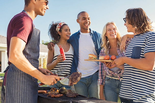 Young multiethnic friends having fun grilling meat enjoying bbq party. Group of happy guys and girls cooking and eating at barbecue dinner outdoor. Men and women standing around grill, chatting, drinking and eating. - Foto, afbeelding