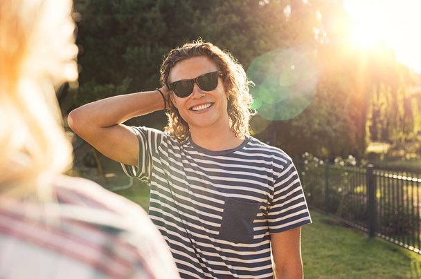 Portrait of happy young man wearing black sunglasses at sunset park. Handsome guy with blonde curly hair looking his girlfriend outdoor. Happy smiling man enjoying the summer. - Φωτογραφία, εικόνα