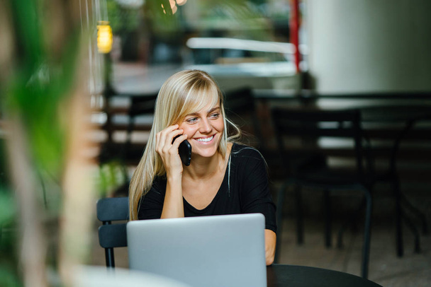 Portrait of a young, attractive and photogenic blond woman talking on her phone with her laptop computer in front of her. She is seated in a cafe during the day as she takes her call. - Foto, Imagen