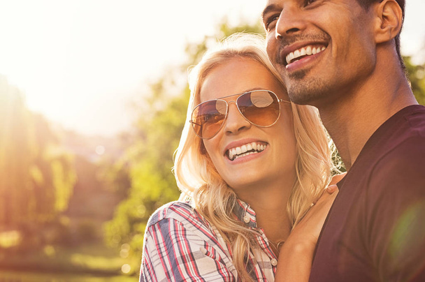 Beautiful young couple hugging and looking away while standing outdoors at sunset. Portrait of happy boyfriend embracing girlfriend with sunglasses at park. Smiling girl hugging guy in love with copy space. - Photo, image