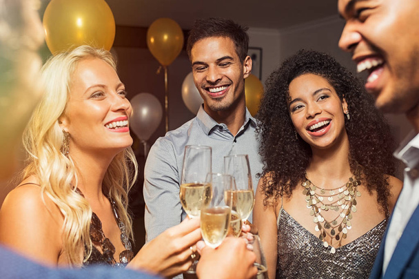 Ομάδα πέντε φίλων πολυεθνική απολαμβάνοντας στο πάρτυ στο σπίτι. Νεαροί άνδρες και όμορφες γυναίκες κατέχουν ποτήρια κρασιού αύξηση τοστ. Χαρούμενα και χαμογελαστά κορίτσια και guys γιορτάζει στο σπίτι με σαμπάνια. - Φωτογραφία, εικόνα