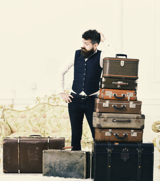Macho elegant on tired face, exhausted at end of packing, leans on pile of vintage suitcases. Man with beard and mustache packed luggage, white interior background. Baggage and relocation concept - Fotoğraf, Görsel