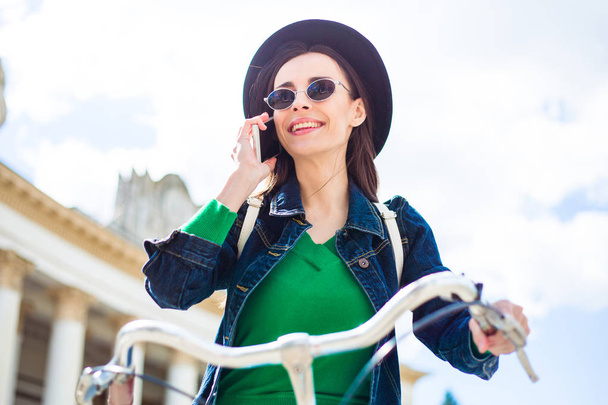 νεαρή γυναίκα στο μαύρο καπέλο να μιλάμε σε smartphone, ενώ ιππασία ποδήλατο εξωτερική - Φωτογραφία, εικόνα