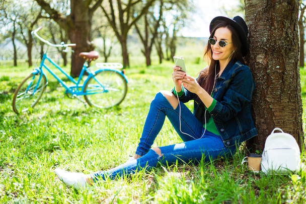 Casual woman in sunglasses and hat sitting on grass and listening music - Photo, image