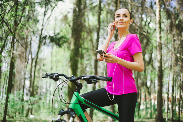 Sporty woman with phone and mountain bicycle in forest - Fotografie, Obrázek
