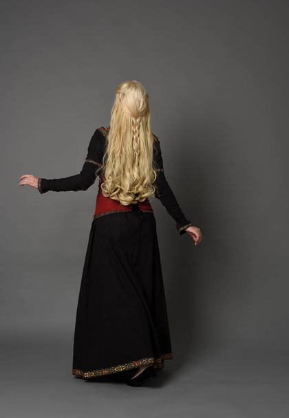 full length portrait of blonde girl wearing red and black medieval costume, standing pose facing away from the camera. - Photo, Image