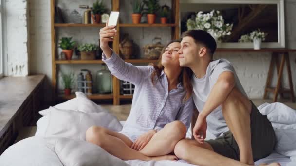 Cheerful loving couple is taking selfie with smartphone looking at camera, posing and making funny faces while sitting together on bed at home. Technology and relationship concept. - Video
