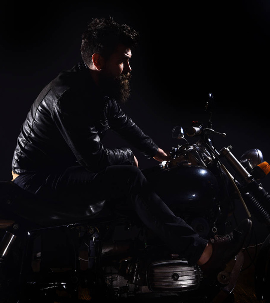 Macho, brutal biker in leather jacket riding motorcycle at night time, copy space. Bikers leisure concept. Man with beard, biker in leather jacket sitting on motor bike in darkness, black background - Photo, Image