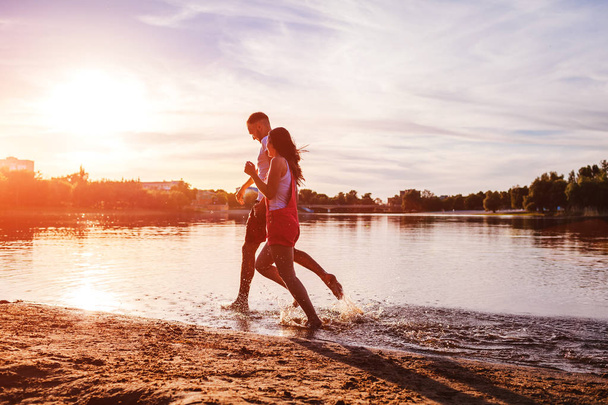 Jovem e mulher correndo na margem do rio de verão. Casal apaixonado se divertindo ao pôr do sol. Rapazes a arrefecer e a desfrutar da liberdade
 - Foto, Imagem
