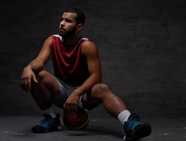 Young African-American basketball player in sportswear sitting on a ball on a dark background. - Φωτογραφία, εικόνα