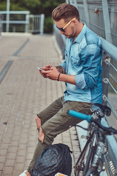 Όμορφος hipster με μοντέρνο κούρεμα στα γυαλιά ηλίου που αναπαύεται μετά την ιππασία σε ένα ποδήλατο, χρησιμοποιώντας ένα smartphone. - Φωτογραφία, εικόνα