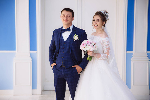 The newlyweds are going to enter the ceremony hall, smiling, the Man in a strict stylish suit and the girl in a white fluffy dress with a bouquet. - Foto, imagen
