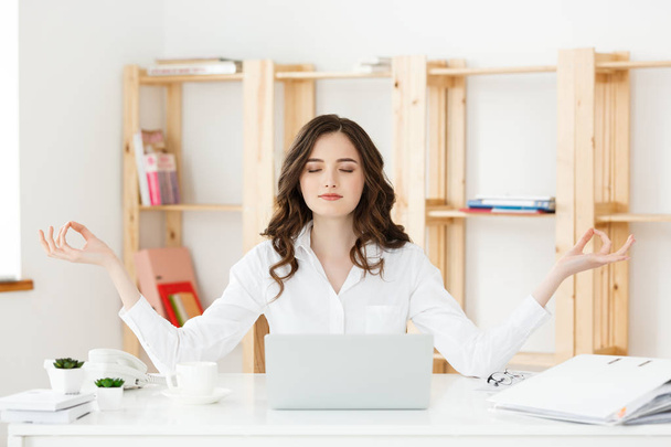 Бизнес и здоровье: Портрет молодой женщины возле ноутбука, практикующей медиацию за офисным столом, перед ноутбуком, онлайн-уроки йоги, делающей перерыв на минуту
. - Фото, изображение