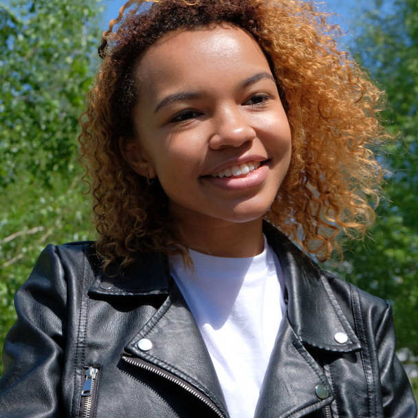 Подростковая мулатка в белой футболке и черная кожаная куртка естественно улыбаются. Портрет красивой чернокожей афроамериканской девушки на улице на фоне зеленых деревьев. Квадратная картина
. - Фото, изображение