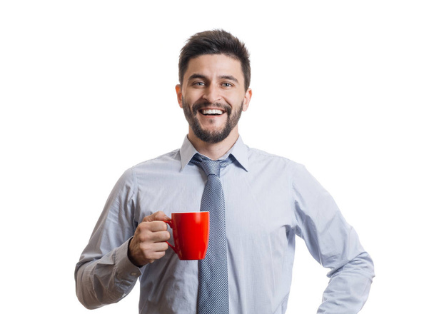 Young cheerful businessman in blue classic shirt smiling with wide opened mouth and holding red mug with drink. Portrait of successful entrepreneur against white background - Фото, изображение
