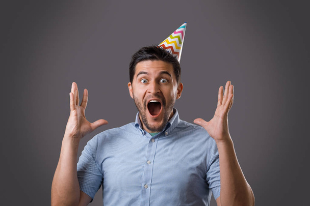 Handsome shocked young brunet guy with party cap screaming with raised hands. New year or birthday celebration - Foto, imagen