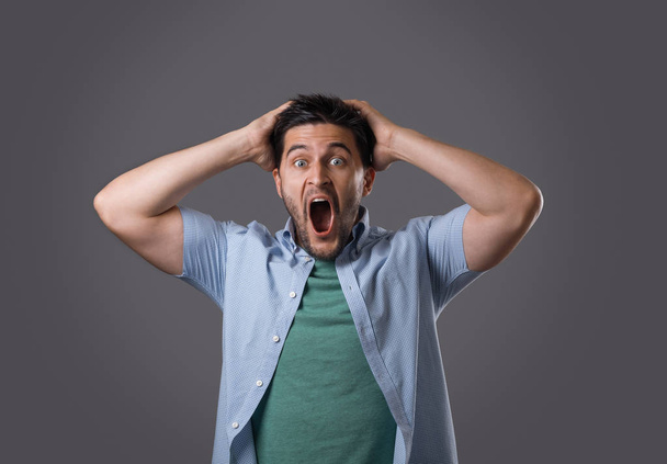 Portrait of young amazed guy that holding his head in hands and yelling isolated on grey background. Student shocked after knowing results of exam - Foto, immagini