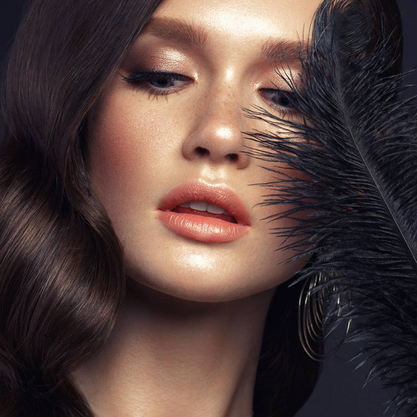 A beautiful girl with evening make-up, a Hollywood wave and a black feather in her hand. Beauty face. - Фото, зображення