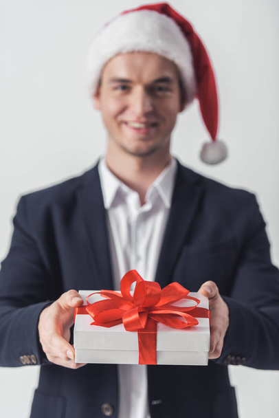 Chico guapo en traje y sombrero de Santa está sosteniendo una caja de regalo, mirando a la cámara y sonriendo, aislado en blanco
 - Foto, imagen