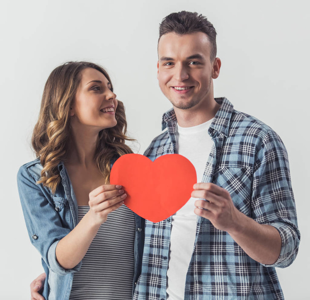 Bella giovane coppia in abiti casual sta tenendo un cuore di carta rossa e sorridente, isolato su bianco
 - Foto, immagini