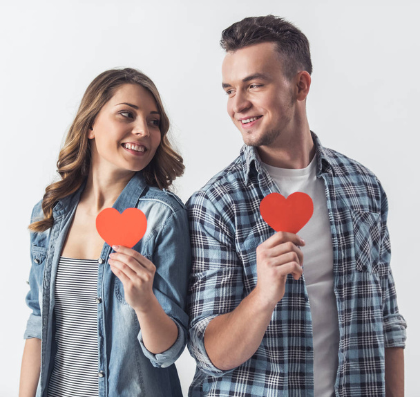 Beautiful young couple in casual clothes is holding two red paper heart, looking at each other and smiling, isolated on white - Fotoğraf, Görsel