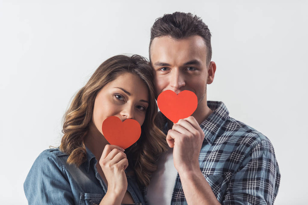Hermosa pareja joven en ropa casual está sosteniendo dos corazones de papel rojo, mirando a la cámara y sonriendo, aislados en blanco
 - Foto, imagen