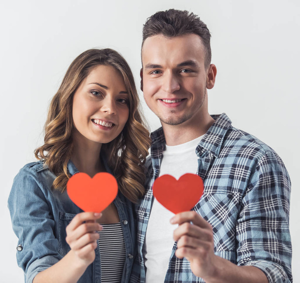 Beautiful young couple in casual clothes is holding two red paper heart, looking at camera and smiling, isolated on white - Photo, Image