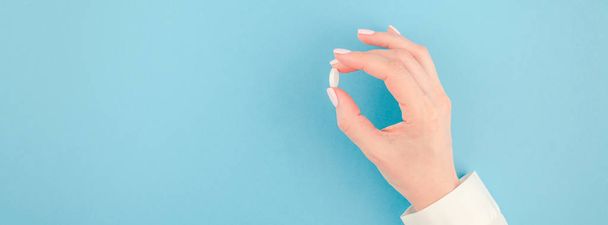 Frau hält eine weiße Pille auf pastellblauem Papierhintergrund mit Kopierraum in minimalistischem Stil, Vorlage für Text. Konzept der medizinischen Behandlung, Pharmazie, Gesundheitsfürsorge. lange breite Fahne - Foto, Bild