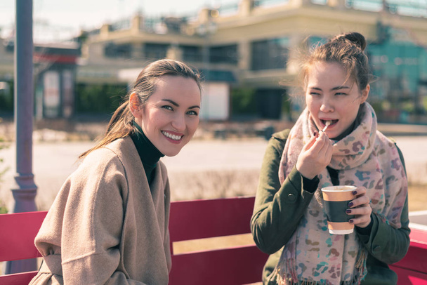 Dos mujeres jóvenes se sientan en un banco de color rosa brillante y beben café en tazas, disfrutando del clima soleado y la espuma del café
. - Foto, imagen