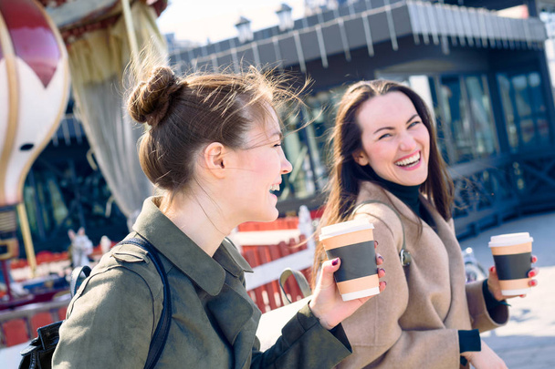 幸せな 2 人の若い女性が大きく微笑し、彼の手でコーヒー カップを保持している晴れた日に街の通りを歩くを笑って. - 写真・画像