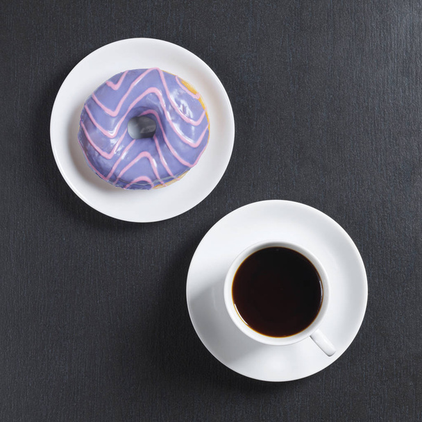 Donut met gekleurde glazuur in witte plaat en kopje koffie op de zwarte stenen achtergrond, bovenaanzicht - Foto, afbeelding