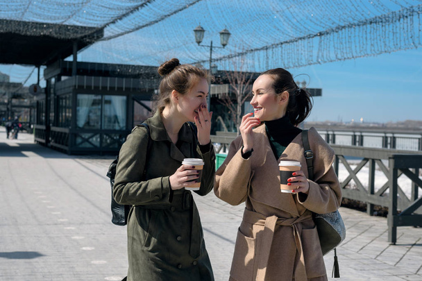 Δύο κορίτσια γέλιο στα παλτά και τα φλιτζάνια του καφέ στα χέρια τους, απολαμβάνουν την υποτροφία. - Φωτογραφία, εικόνα