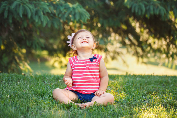 Портрет милой очаровательной улыбающейся маленькой белой малышки в парке снаружи. Концепция счастливого детства. Смеющийся веселый ребенок сидит на траве и смотрит вверх во время летнего заката
 - Фото, изображение