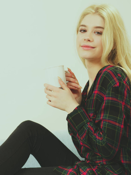 Pretty hipster young adult teenage woman sitting on sofa enjoying her leisure time drinking coffee or tea from mug. - Foto, imagen
