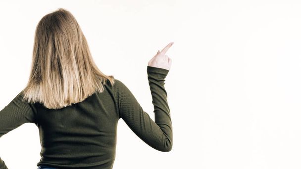 Vista posterior de mujer con cabello rubio sobre fondo blanco mostrando algo, apuntando con el dedo al espacio de copia
. - Foto, imagen