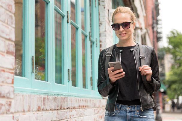 Mulher jovem na moda em jaqueta de couro preto e ocupado com seu telefone móvel enquanto caminha uma rua da cidade
 - Foto, Imagem