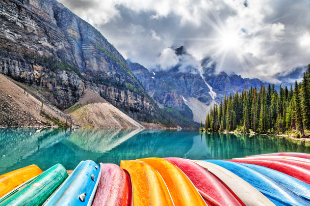 A row of canoes line the shores of Moraine Lake in Lake Louise of the Canadian Rockies, as the sun peeks through the heavy clouds that descended on the Valley of Ten Peaks in the background. - Photo, Image