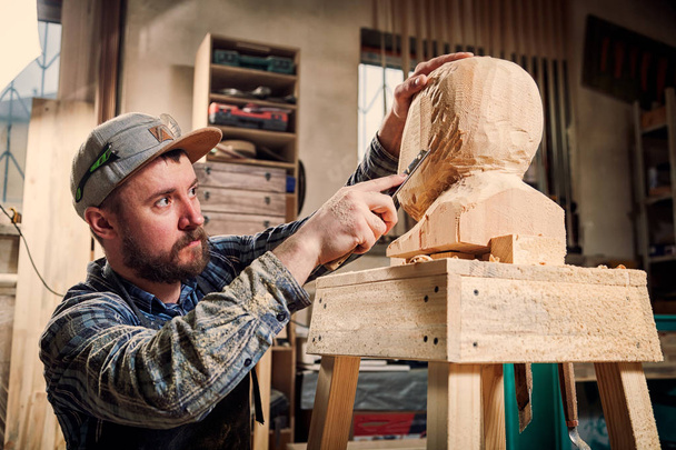 Nuori puuseppä työvaatteissa sahaa miehen pään puulla, käyttäen talttaa työpajassa, noin paljon työkaluja työhön
 - Valokuva, kuva