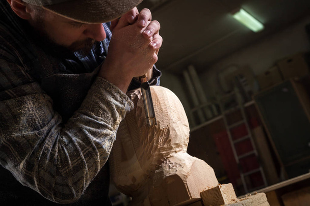 Close up jovem carpinteiro em uma roupa de trabalho serra a cabeça de um homem com uma árvore, usando um cinzel na oficina, em torno de um monte de ferramentas para o trabalho
 - Foto, Imagem
