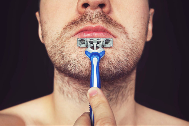 Jeune homme sans crème à raser sur son visage, se toilettant la barbe avec un rasoir droit sur fond noir - Photo, image