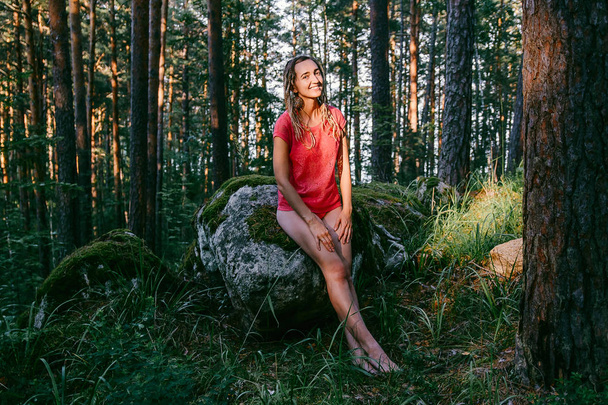 beautiful tanned girl sitting on a big stone and smiles in the woods - Photo, image