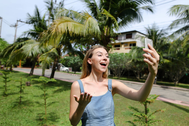 背景のヤシの木の近くの smarthone で selfie を作って笑顔の少女. - 写真・画像